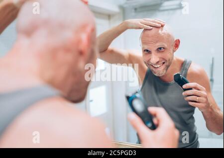 Haarlose Mann zufrieden mit seinem neuen Stil Frisur macht er mit einem elektrischen wiederaufladbaren Trimmer suchen im Badezimmer Spiegel. Radikale Entscheidung, lebenslange Stockfoto