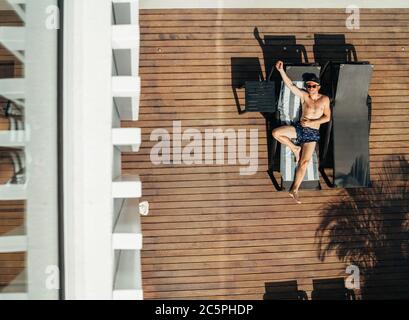 Draufsicht auf Mann liegend und Sonnenbräune auf Liegestuhl gekleidet Badehose, Baseball-Cap und Sonnenbrille. Sorgloser Mann Urlaub Konzept Bild. Stockfoto