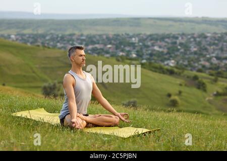 Ein bärtiger, nicht junger Mann praktiziert Yoga-Meditation, sitzt in einer Lotusposition auf dem Gras auf der Sommernatur. Stockfoto