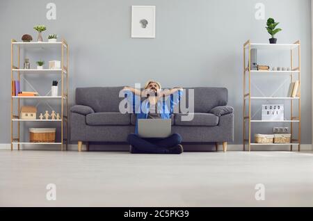 Zufriedener Geschäftsmann in legerer Kleidung mit einem Laptop träumt auf dem Boden im Wohnzimmer sitzen. Stockfoto
