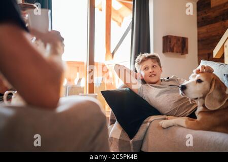 Gemütliche Familie Tee Zeit. Vater und Sohn im Wohnzimmer. Junge auf einem bequemen Sofa liegend und ihren Beagle Hund streichelte und lächelte. Friedliche Familie Stockfoto