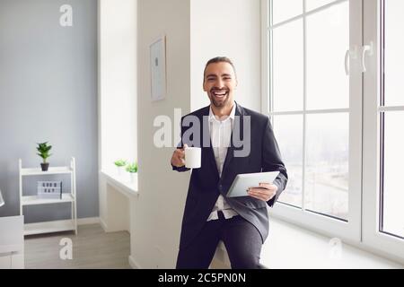 Ein Mann in einer schwarzen Jacke mit einem Kaffeebecher in der Hand lächelt, während er am Fenster im Büro steht Stockfoto