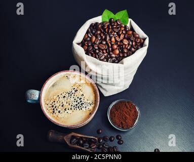 Foto von Kaffeetasse, geröstete Kaffeebohnen in Leinwandbeutel und gemahlenes Pulver auf schwarzem Küchentisch. Stockfoto