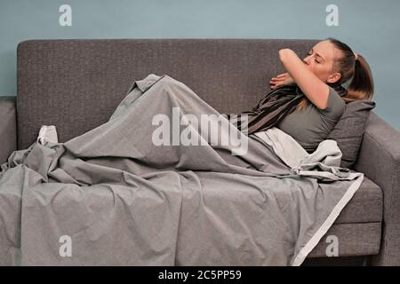 Das Mädchen ist zu Hause krank. Auf einer Couch sitzend, in eine Decke gewickelt, ein Schal um seinen Hals. Hustet am Ellenbogen. Coronavirus. Stockfoto
