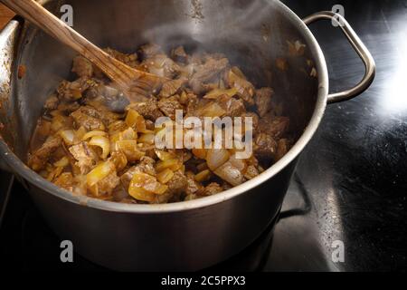 Kochen Dampfgaren von Eintopf, Gulasch oder Ragout mit Rind und Zwiebeln in einem Stahltopf auf einem schwarzen Herd, ausgewählter Fokus, enge Schärfentiefe Stockfoto