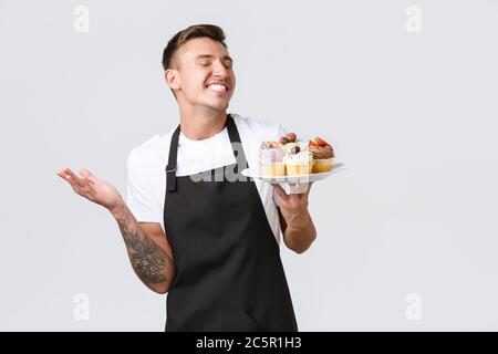 Kleineinzelhandel, Cafés und Bäckereikonzept. Lächelnd, gut aussehender Barista, Kellner im Laden, der Teller mit köstlichen Desserts hält und serviert Stockfoto