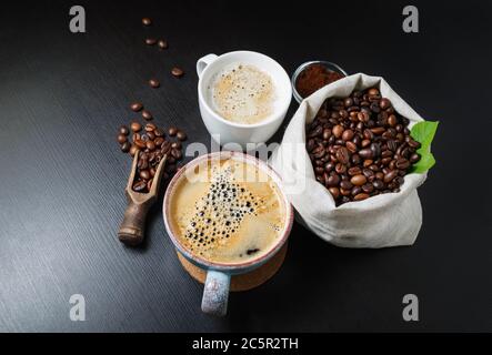 Stillleben mit Kaffeetassen und Kaffeebohnen in Leinenbeutel auf schwarzem Küchentisch. Stockfoto