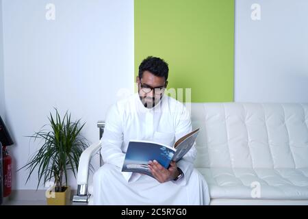 Saudi man sitzt im Wartebereich im Büro auf Couch/Sofa, während man ein Magazin liest Stockfoto