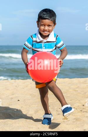 Ein Sri-lankischer Junge spielt am frühen Morgen mit einer roten Plastikkugel am Strand von Negombo in Sri Lanka. Stockfoto