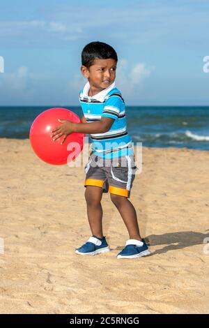 Ein Sri-lankischer Junge spielt am frühen Morgen mit einer roten Plastikkugel am Strand von Negombo in Sri Lanka. Stockfoto