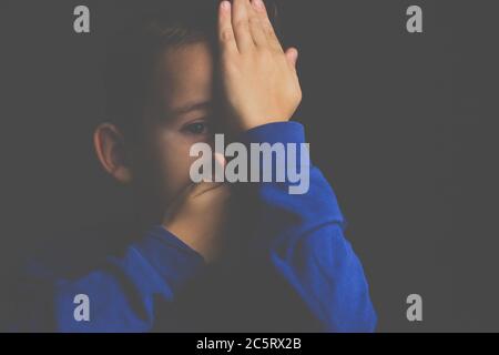 Porträt eines dunkelhaarigen Jungen, der sein Gesicht mit den Händen auf einem dunklen Hintergrund bedeckt Stockfoto