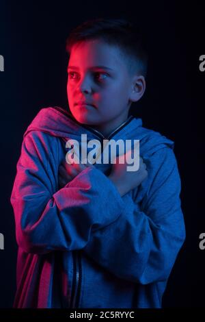 Studio-Portrait eines dunkelhaarigen Jungen, auf dunklem Hintergrund, in einer Kapuzenjacke in der Kapuze. Helle Farbe: Rot und blau Stockfoto