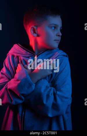 Studio-Portrait eines dunkelhaarigen Jungen, auf dunklem Hintergrund, in einer Kapuzenjacke in der Kapuze. Helle Farbe: Rot und blau Stockfoto