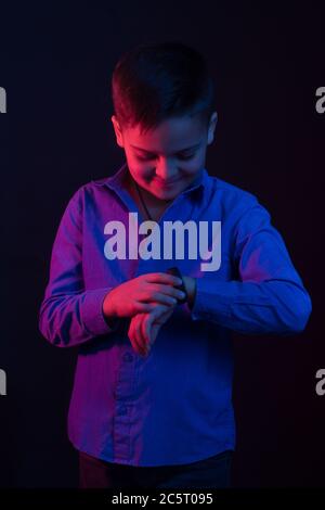 Studio-Portrait eines dunkelhaarigen Jungen, auf dunklem Hintergrund, in einer Kapuzenjacke in der Kapuze. Helle Farbe: Rot und blau, er sieht auf seine Uhr Stockfoto