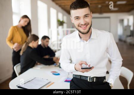 Junger lächelnder Mann mit Bart in weißem Hemd, der fröhlich in die Kamera schaut und das Handy in der Hand hält, während er Zeit im Büro mit Kollegen verbringt Stockfoto