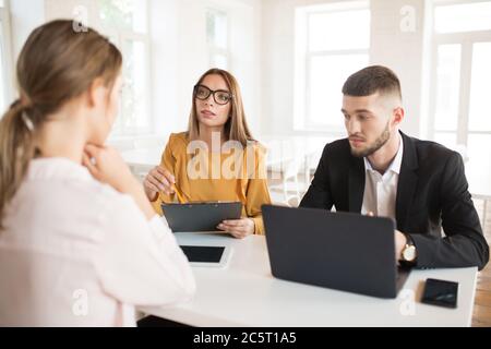 Business-Mann mit Laptop nachdenklich beiseite schauen, während Business-Frau in Brillen mit Ordner in der Hand im Gespräch mit Bewerber über die Arbeit. Jung Stockfoto