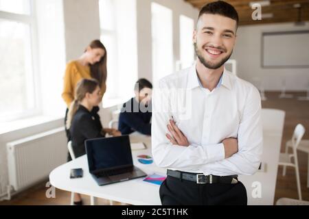 Junger lächelnder Mann mit Bart in weißem Hemd, der mit gefalteten Händen fröhlich in die Kamera schaut, während er Zeit im Büro mit Kollegen auf dem Hintergrund verbringt Stockfoto