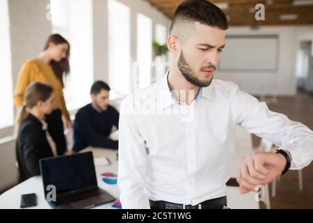 Junger Mann mit Bart in weißem Hemd nachdenklich Blick auf Handuhr, während Zeit im Büro mit Kollegen auf Hintergrund Stockfoto