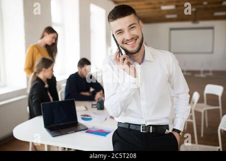 Junger lächelnder Mann mit Bart in weißem Hemd, der fröhlich in die Kamera schaut und auf dem Handy spricht, während er Zeit im Büro mit Kollegen verbringt Stockfoto