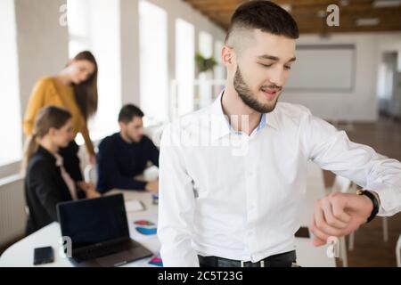 Junger Mann mit Bart in weißem Hemd auf Handuhr schauen, während Zeit im Büro mit Kollegen auf Hintergrund verbringen Stockfoto