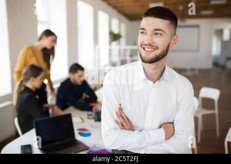 Junger lächelnder Mann mit Bart in weißem Hemd, der mit gefalteten Händen glücklich zur Seite schaut, während er Zeit im Büro mit Kollegen auf dem Hintergrund verbringt Stockfoto