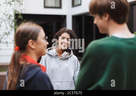 Fröhliche Dame mit dunklen lockigen Haaren in Kopfhörer stehend und glücklich mit ihren Freunden sprechen, während sie Zeit im Hof der Universität zu verbringen Stockfoto