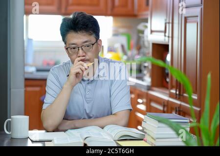 Asiatischer Mann mittleren Alters, der zu Hause am Schreibtisch sitzt, ein Buch liest und studiert. Stockfoto