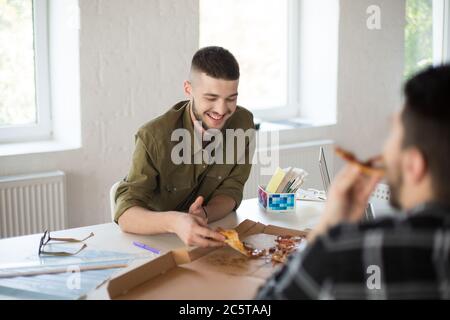 Junger lächelnder Mann mit Bart im Hemd, der fröhlich Pizza isst und Zeit im modernen Büro verbringt Stockfoto