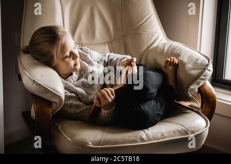 Ein blondes kaukasisches Mädchen sitzt zusammengerollt auf einem Stuhl ein Buch zu lesen Stockfoto