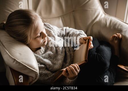 Ein blondes kaukasisches Mädchen sitzt zusammengerollt auf einem Stuhl ein Buch lesen Stockfoto