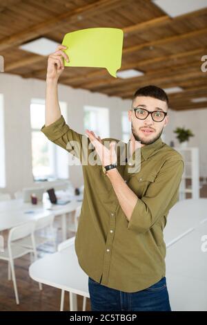 Junger Mann in Brille und Hemd erstaunt in der Kamera mit gelben Papier Form der Nachricht unter dem Kopf, während die Zeit bei der Arbeit in modernen Stockfoto