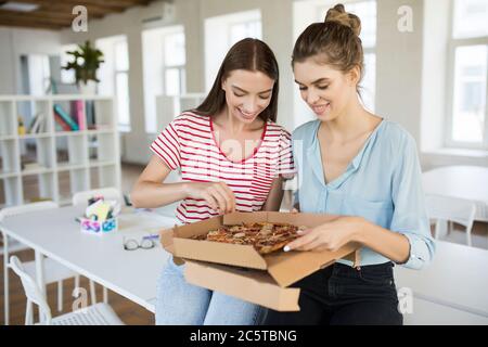 Schöne lächelnde Mädchen sitzen auf der Rezeption glücklich Pizza essen, während Zeit zusammen in modernen Büro verbringen Stockfoto