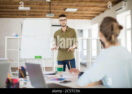Junger Mann in Brille und Hemd neben Bord stehend nachdenklich über neues Projekt mit Kollegen reden. Gruppe von kreativen Menschen arbeiten Stockfoto