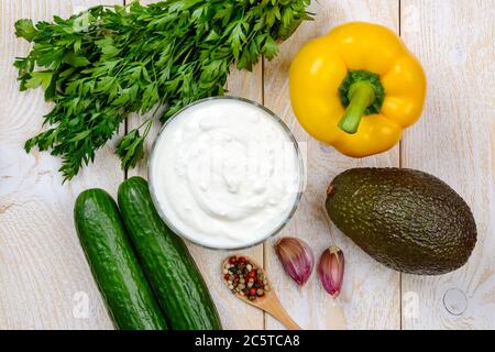 Zutaten für rohes Gurkensuppenpüree: griechischer Joghurt, Gurke, Avocado, gelber Paprika, Petersilie und Knoblauch auf einem Holztisch. Rohkost. Stockfoto