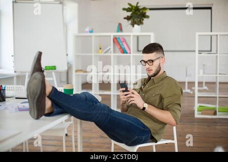 Junger Mann in Brille und Hemd mit Handy, während auf Stuhl mit Beinen auf dem Tisch bei der Arbeit sitzen. Durchdachte Geschäftsmann, der Zeit in modernen verbringen Stockfoto