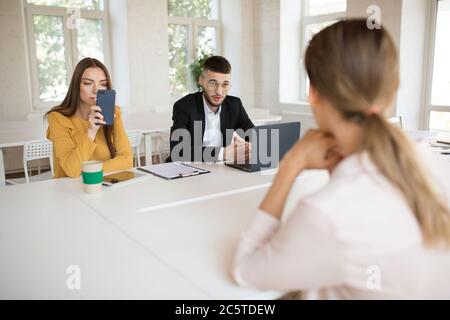 Junge Geschäftsmann in Brillen mit Laptop im Gespräch mit Bewerber, während Business-Frau nachdenklich mit Handy. Junge Arbeitgeber, die Arbeit ausgeben Stockfoto
