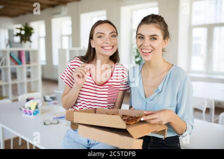 Zwei fröhliche Mädchen sitzen auf dem Schreibtisch mit Pizza glücklich Blick in die Kamera, während die Zeit zusammen in modernen Büro Stockfoto