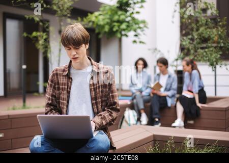 Foto von verärgert jungen Mann auf der Bank sitzen und leider auf Laptop arbeiten, während die Zeit im Hof der Universität mit Studenten auf dem Hintergrund Stockfoto
