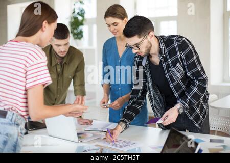 Nachdenklicher Mann in Hemd und Brille, der mit Skizzen arbeitet, während er Zeit mit Kollegen bei der Arbeit verbringt. Gruppe junger kreativer Menschen, die in arbeiten Stockfoto