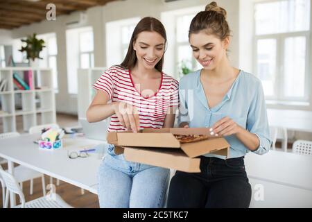 Zwei hübsche Mädchen sitzen gerne auf dem Schreibtisch mit Pizza, während sie Zeit zusammen im modernen Büro verbringen Stockfoto
