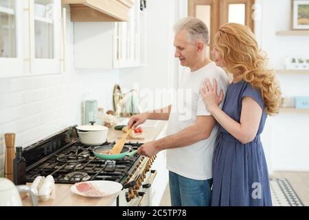 Seitenansicht Porträt von reifen Paar Kochen zusammen in der Küche mit eleganten Frau umarmen Mann von hinten, kopieren Raum Stockfoto