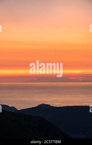 Sonnenuntergang über dem ruhigen Mittelmeer Meer mit der Silhouette des Hügeldorfes Sant'Antonino auf Korsika im Vordergrund und Küste von Frankreich in Stockfoto