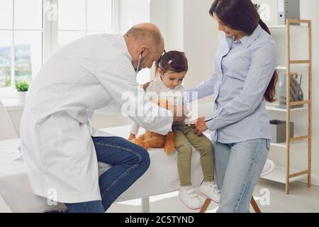 Oberarzt mit Stethoskop hört einem kleinen Mädchen mit Mutterpatienten im Krankenhaus zu. Stockfoto