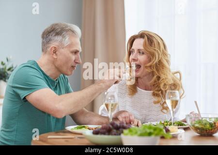 Portrait von modernen reifen Paar genießen Abendessen zusammen zu Hause, Mann gibt Frau Stück Essen auf Gabel, kopieren Raum Stockfoto