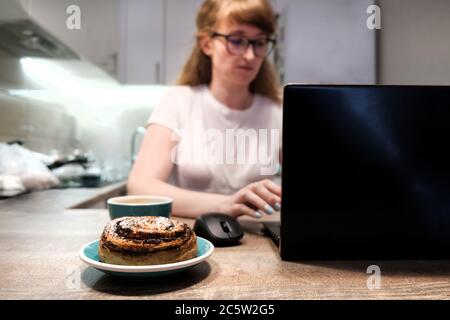Konzentrierte junge Frau mit Brille zu Hause mit Laptop in der Küche sitzen. Sie ist so beschäftigt, dass sie keine Zeit hat, ihren Morni zu trinken Stockfoto
