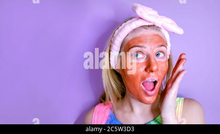 Pflege der Haut zu Hause. Junge Frau mit einer Tonmaske auf ihrem Gesicht und Haarband auf dem Kopf berühren ihre Haut sanft Stockfoto
