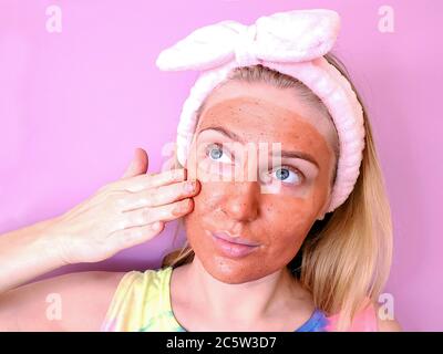 Pflege der Haut zu Hause. Junge Frau mit einer Tonmaske auf ihrem Gesicht und Haarband auf dem Kopf berühren ihre Haut sanft Stockfoto