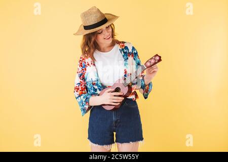 Portrait einer jungen hübschen Dame in Strohhut und buntem Hemd, das mit kleiner Gitarre steht und auf dem sie auf rosa Hintergrund fröhlich spielt Stockfoto