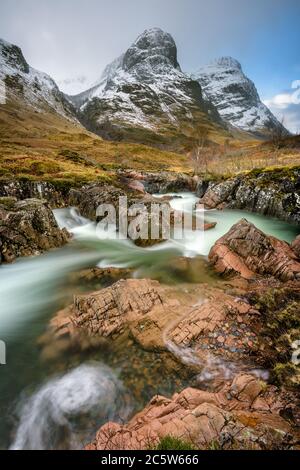 Die Bergkette der Three Sisters in Glencoe ist mit Winterschnee bedeckt und im Vordergrund ein interessanter felsiger Bach. Schottische Highlands, Großbritannien. Stockfoto