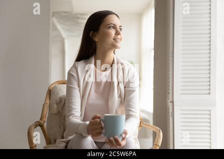 Lächelnd verträumte junge Frau hält Tasse, sitzt in gemütlichen Sessel Stockfoto